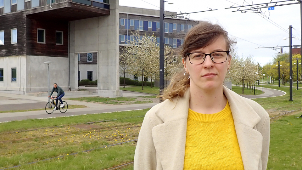 woman in yellow sweater stands outside a building with a tramway in the background. Photo. 