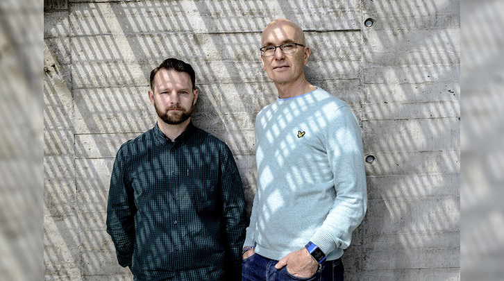 Two men in front of a concrete wall. Photo. 